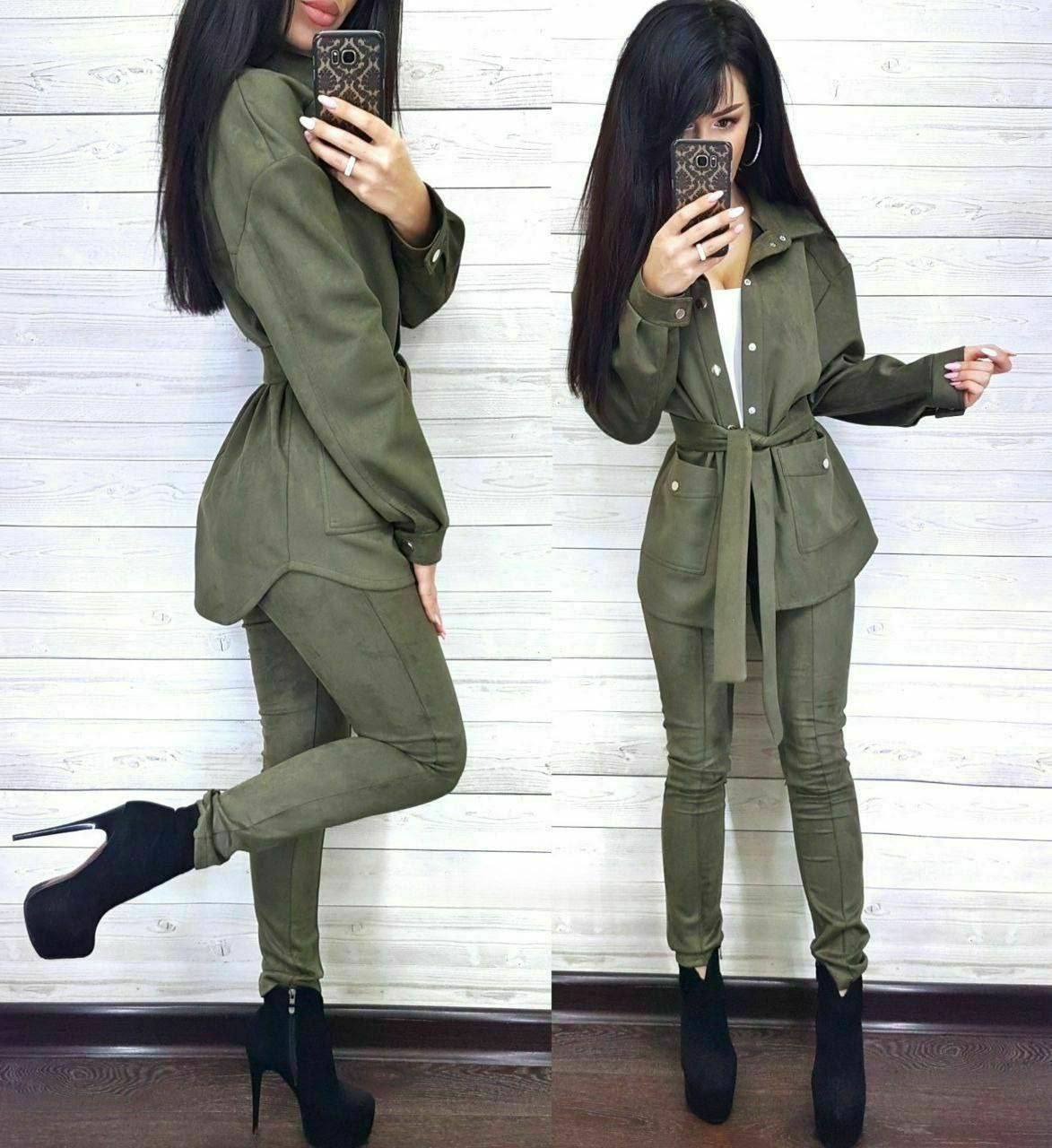 Woman wearing a green outfit with a belt, standing against a white wall.