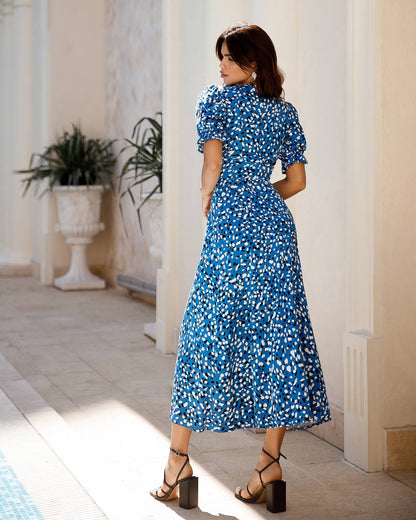 Woman in a blue floral dress standing in a sunlit room with columns and plants.