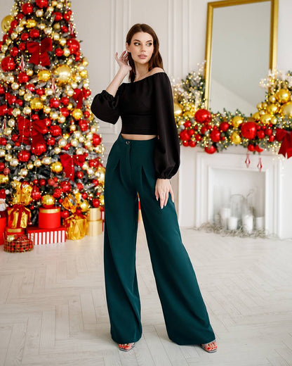 Woman in black top and green pants standing in a decorated room with Christmas tree and fireplace.