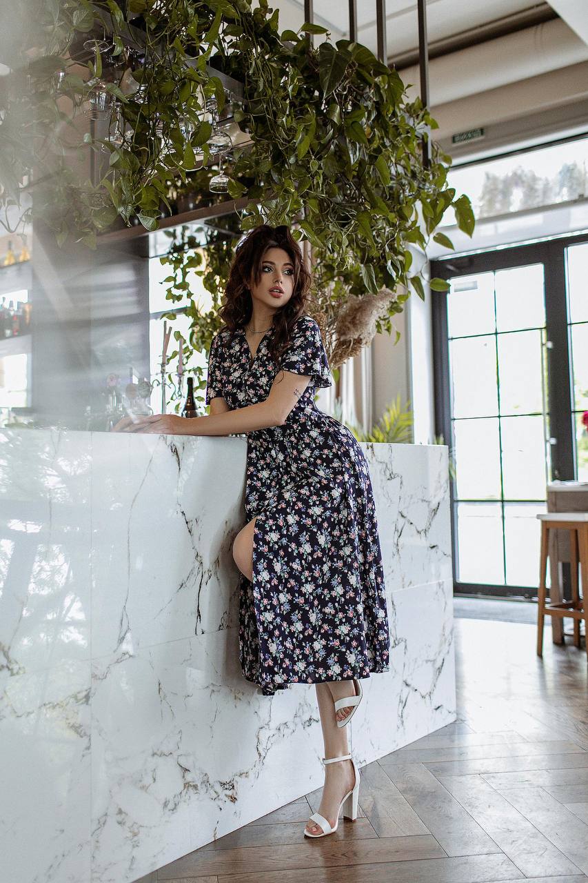 Woman in a floral dress leaning against a marble wall with plants above.