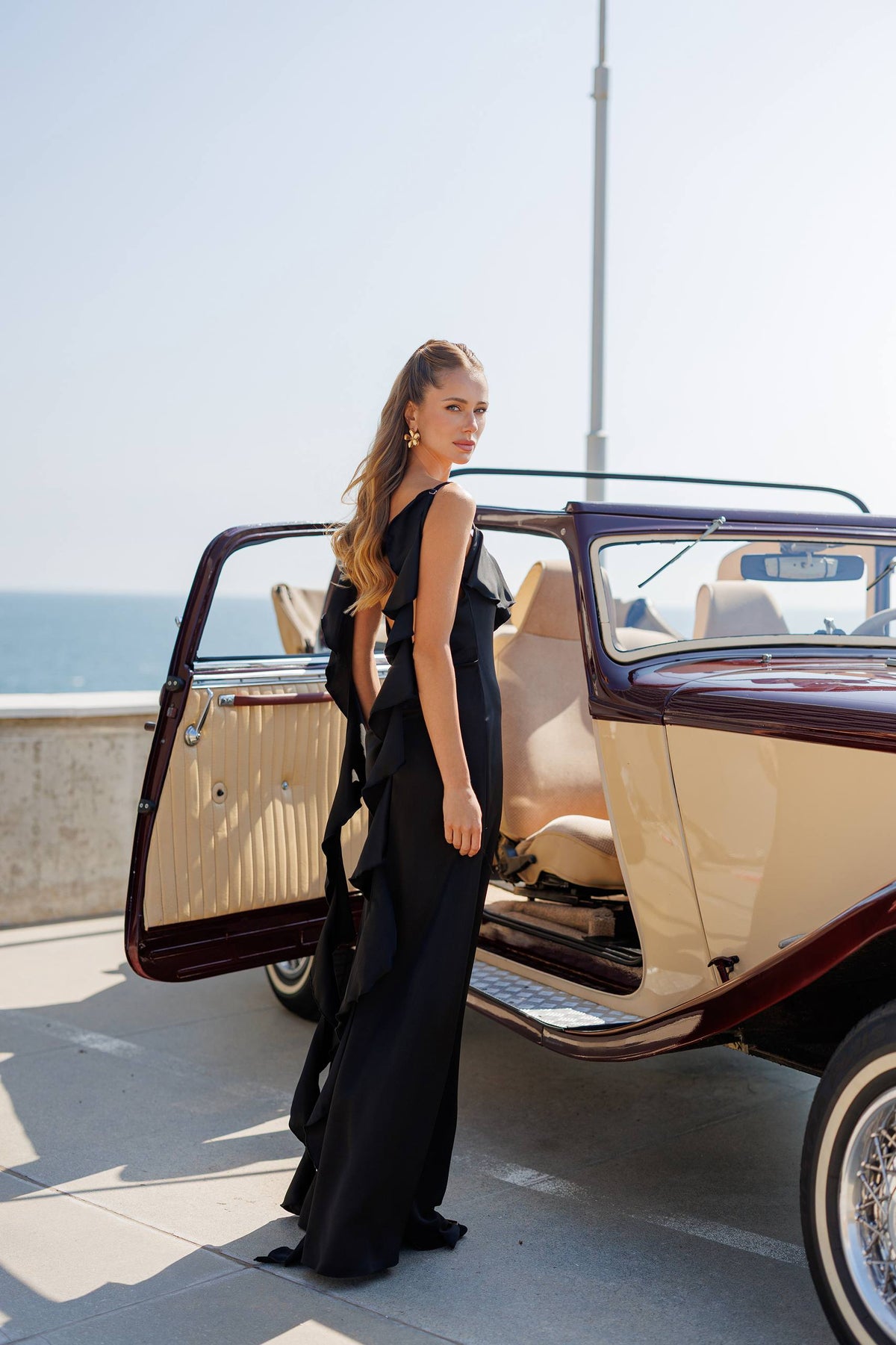 Woman in a black dress standing next to an open vintage car by the sea.