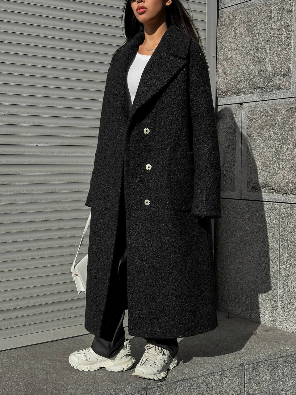 Person wearing a long black coat standing against a gray wall.