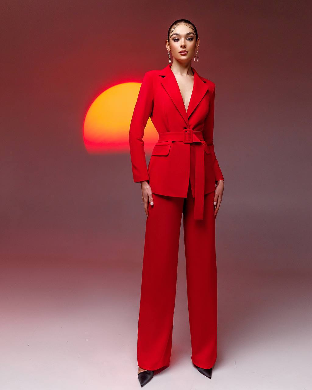 Woman in a red suit standing against a gradient background with a sun-like shape.