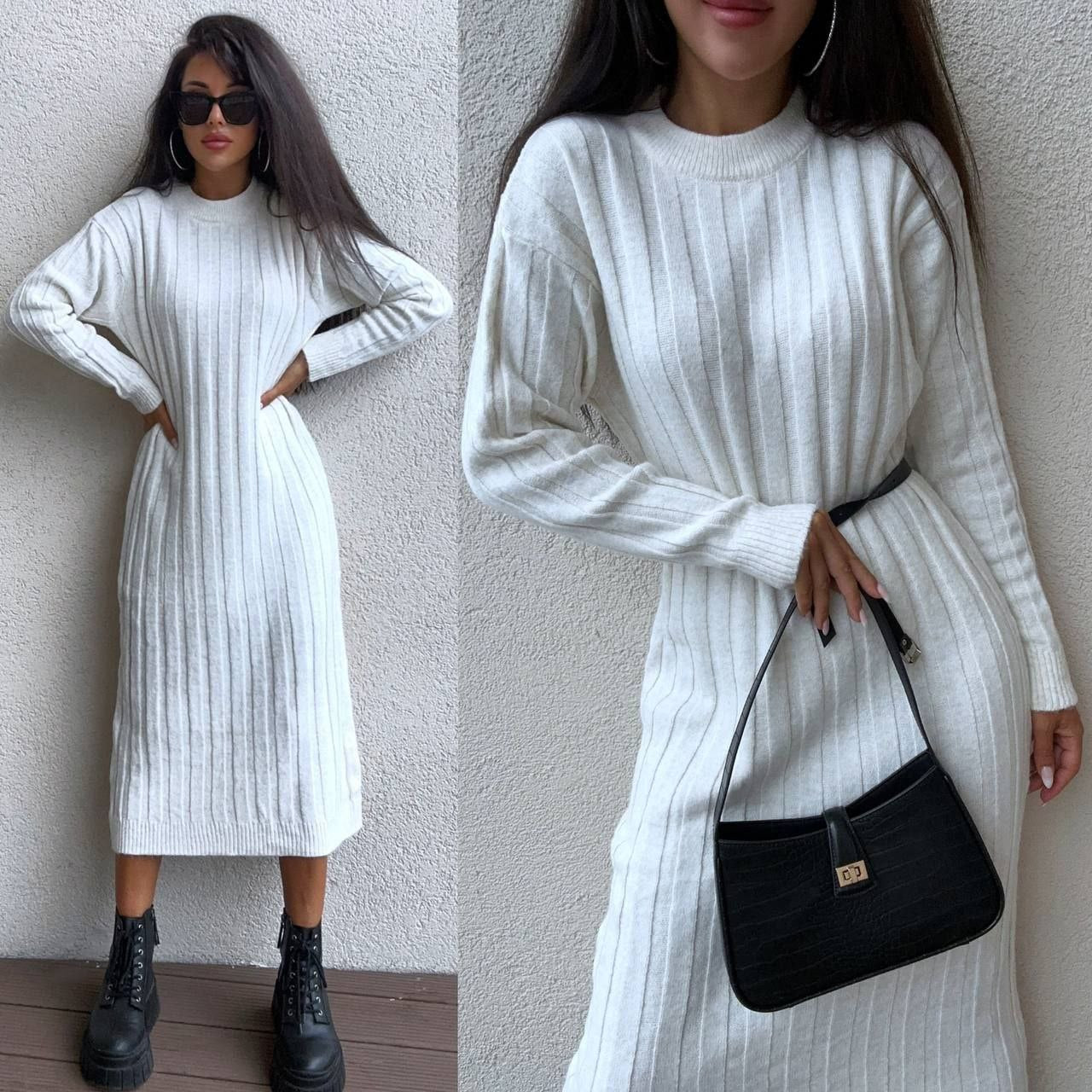 Woman wearing a white ribbed dress with black boots and a black handbag against a plain wall.