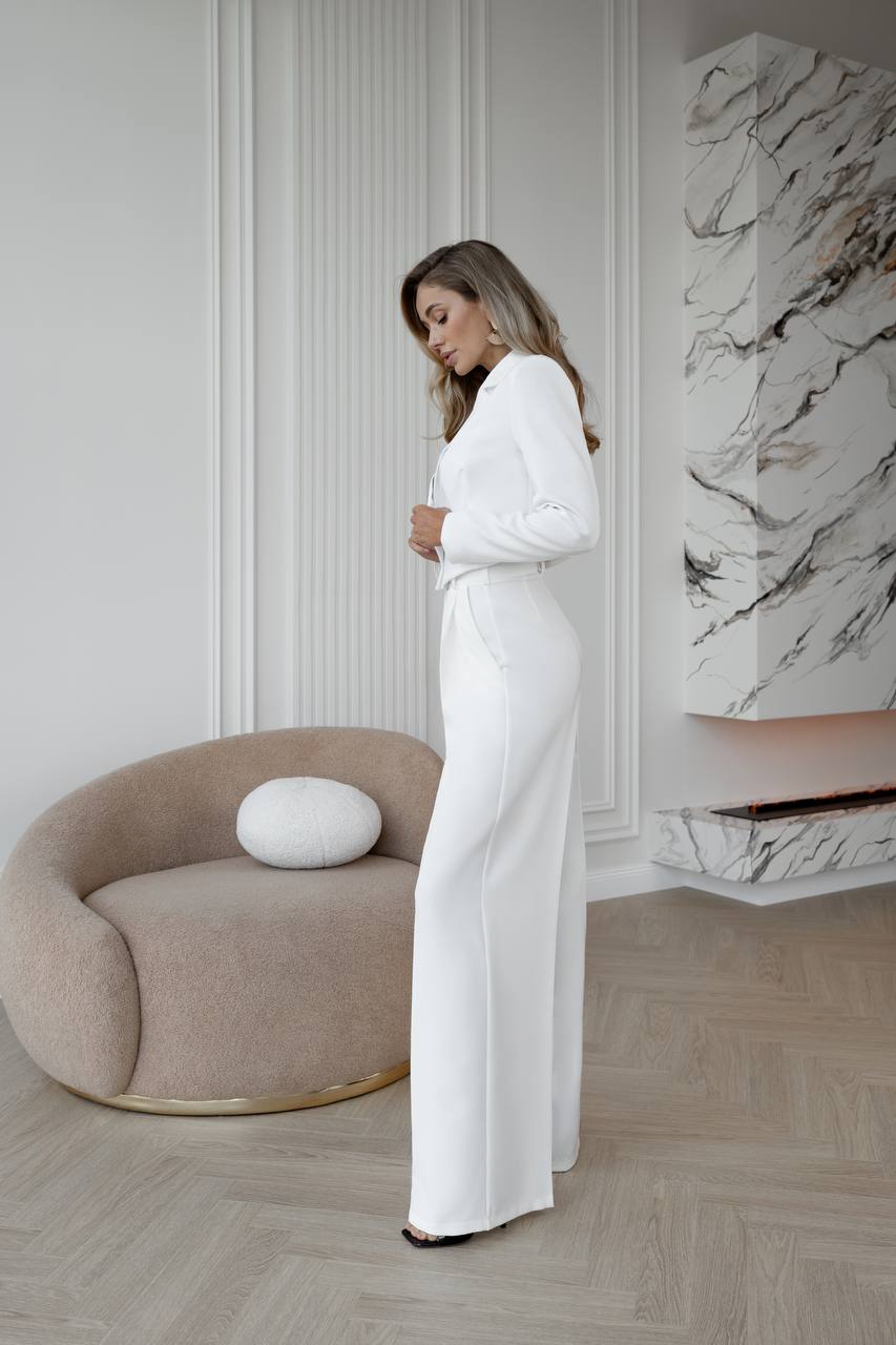 Woman in a white outfit standing in a modern room with a marble wall and beige chair.