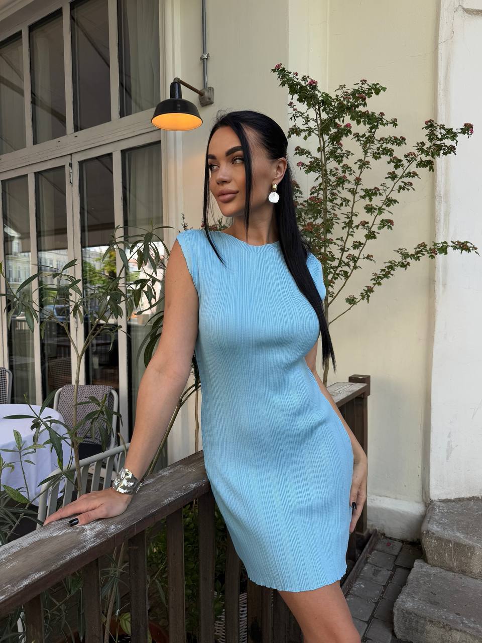 Woman in a light blue dress standing on a balcony with plants and a building in the background.