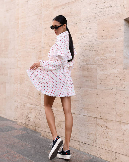 Woman in a white polka dot dress standing against a beige wall.