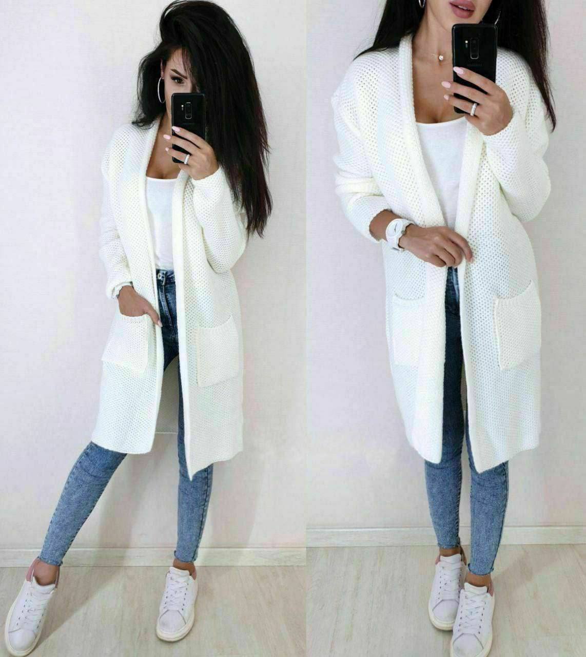 Woman wearing a long white cardigan over a white top and blue jeans, taking a mirror selfie.