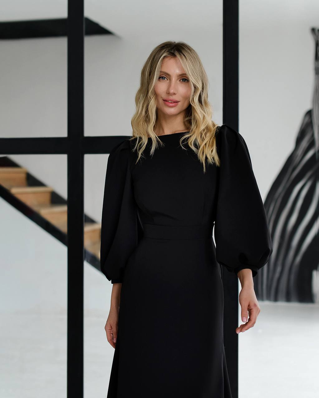 Woman wearing a black dress standing in an indoor setting with a staircase.