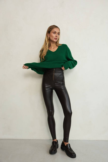 Woman wearing a green sweater and black leather pants against a plain background