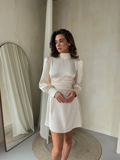Woman in a white dress standing in a room with a mirror and light-colored walls.
