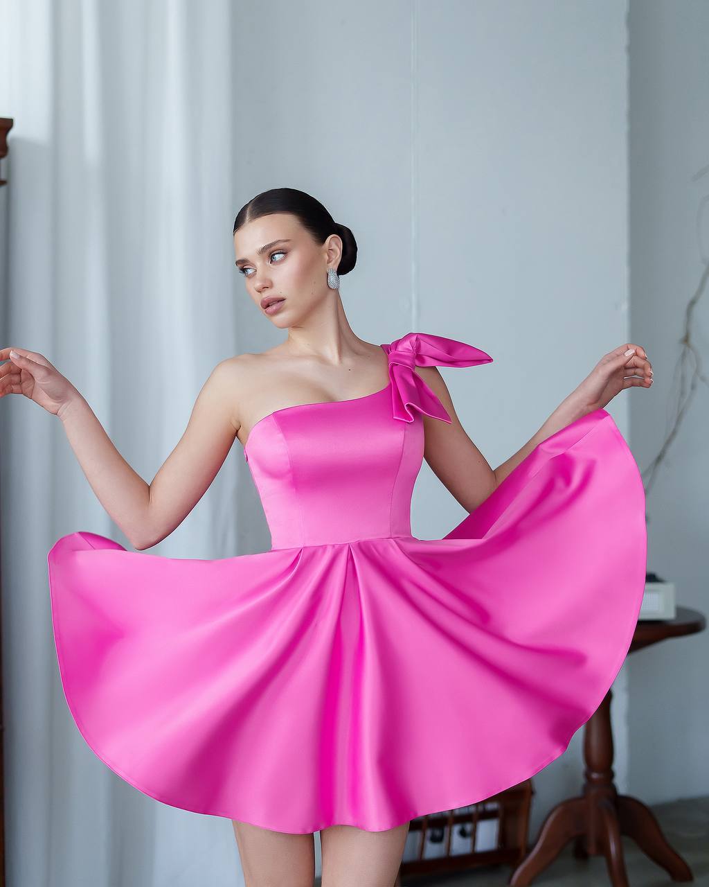 Woman wearing a pink one-shoulder dress with a bow detail, standing indoors.