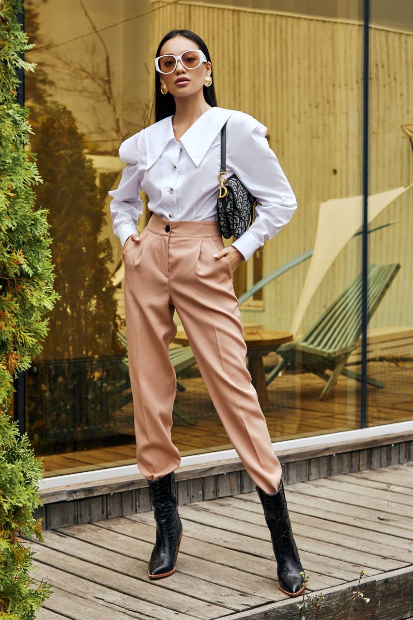 Woman in a white blouse and pink pants standing outdoors near a glass wall.
