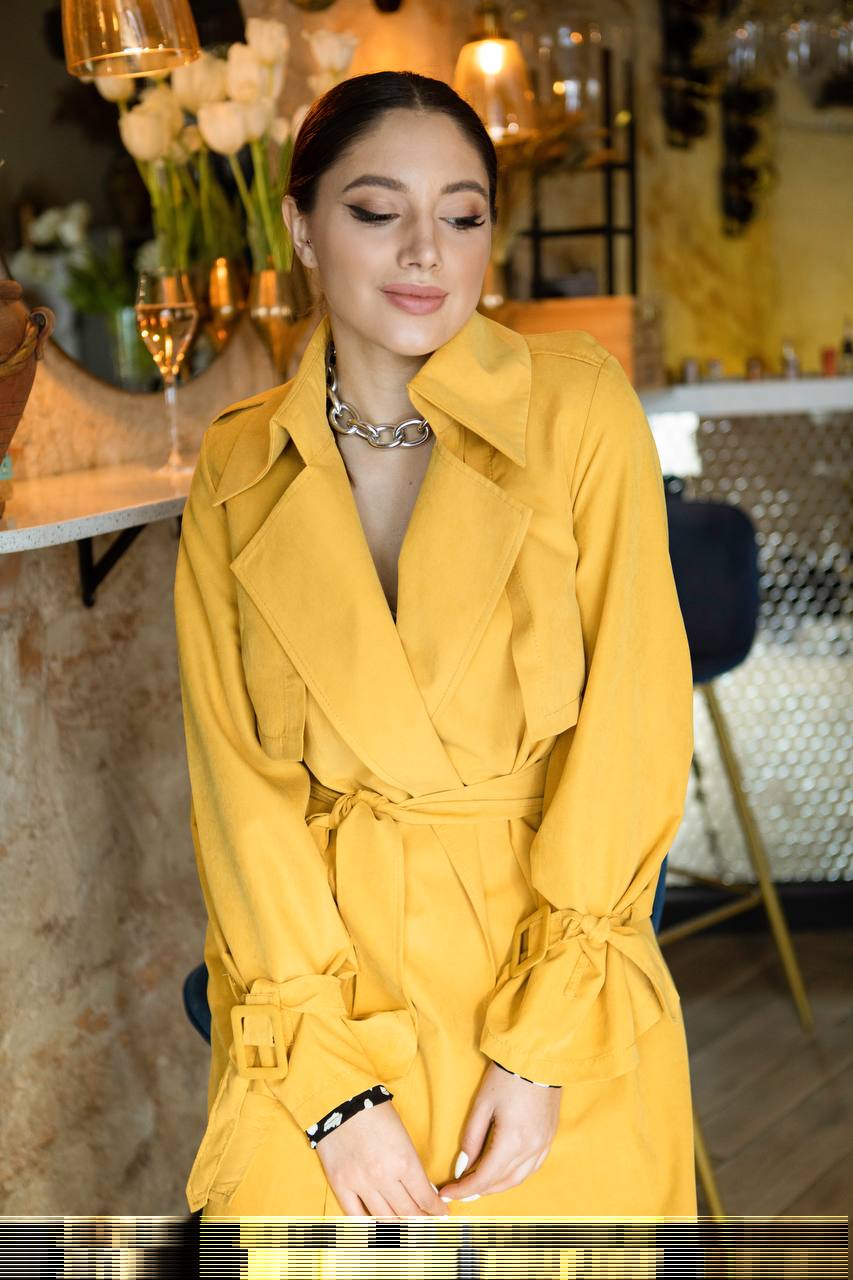 Woman wearing a yellow trench coat in an indoor setting with decor elements.