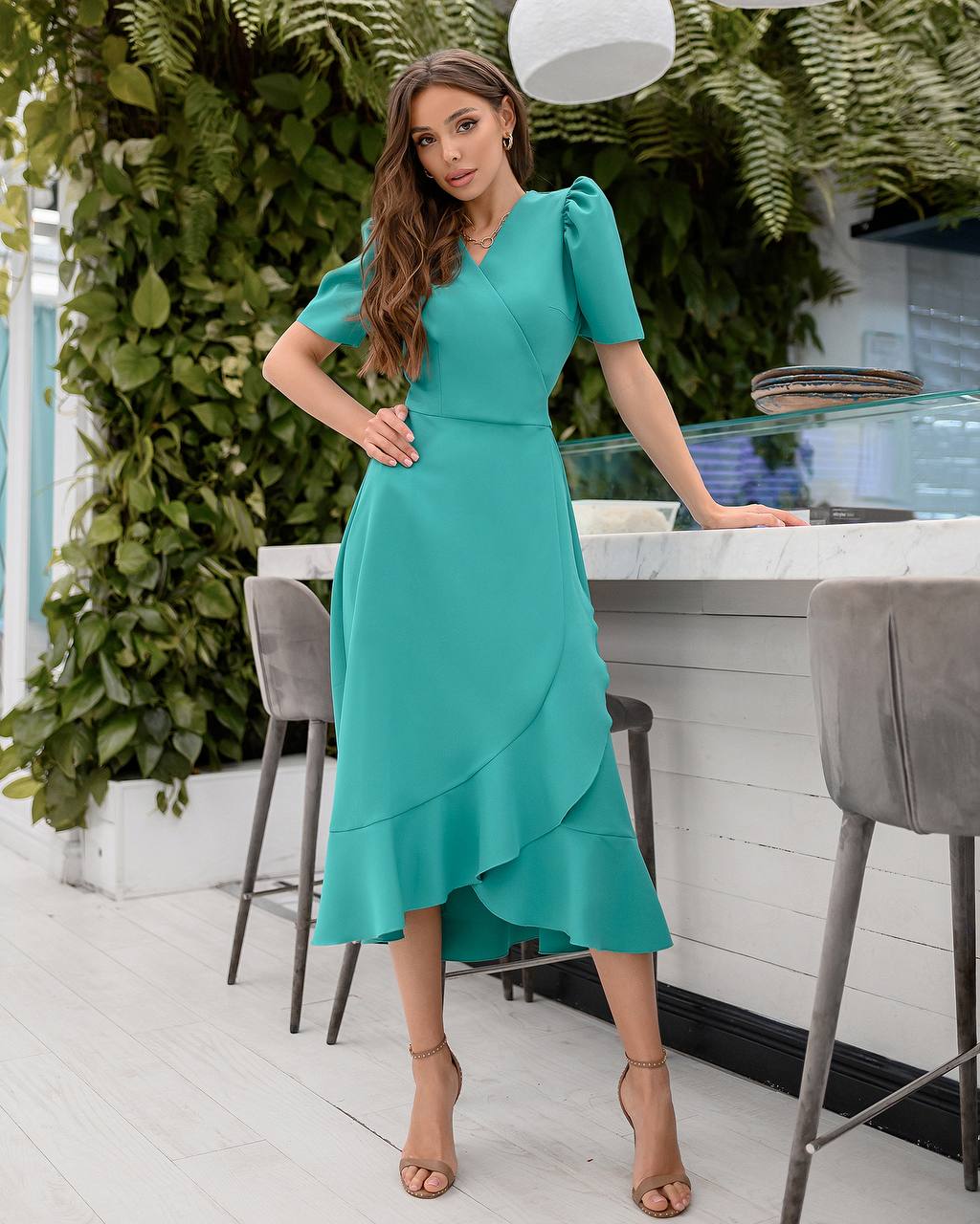 Woman in a teal dress standing in a modern kitchen with greenery.