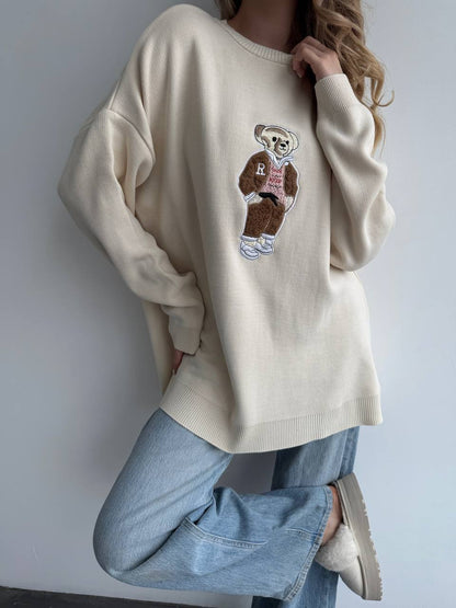 Person wearing a beige sweater with a teddy bear graphic and text, standing against a plain background.