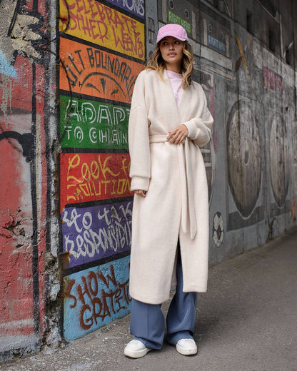 Person wearing a long cream coat standing in front of a colorful graffiti wall.