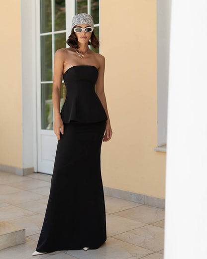 Woman wearing a black strapless dress and sunglasses in front of a beige wall.
