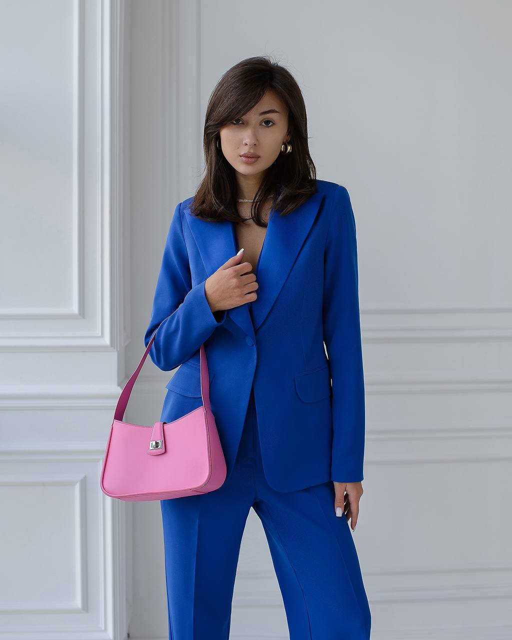 Woman in a blue suit holding a pink handbag against a white wall