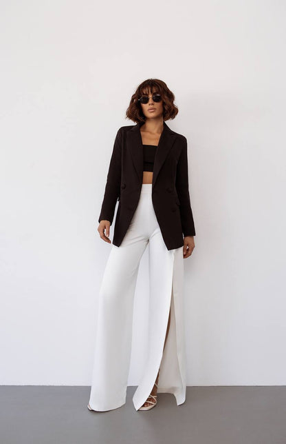 Woman wearing a brown blazer, black top, and white pants against a white background