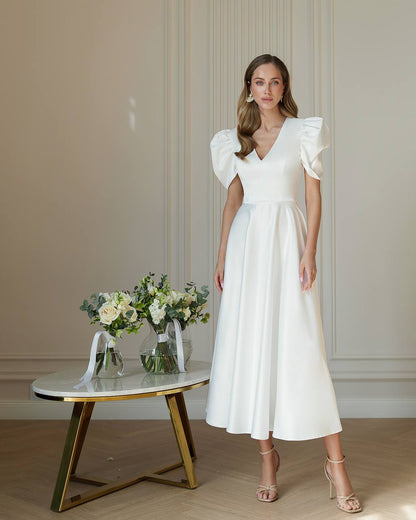 Woman in a white dress standing next to a small table with flowers against a neutral wall.