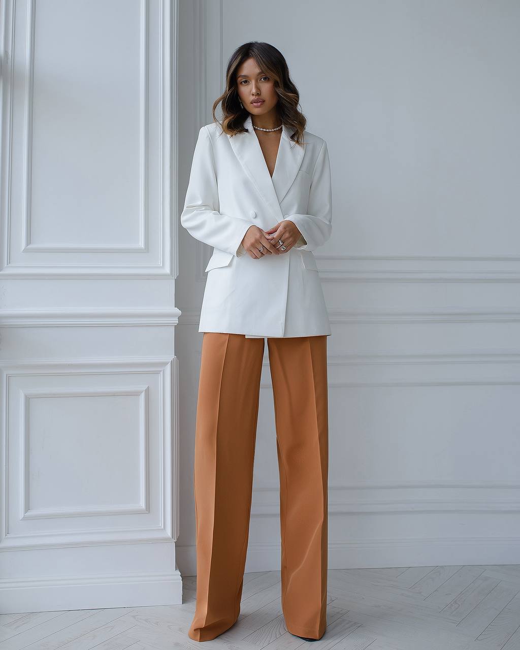 Woman wearing a white blazer and brown pants against a white wall.