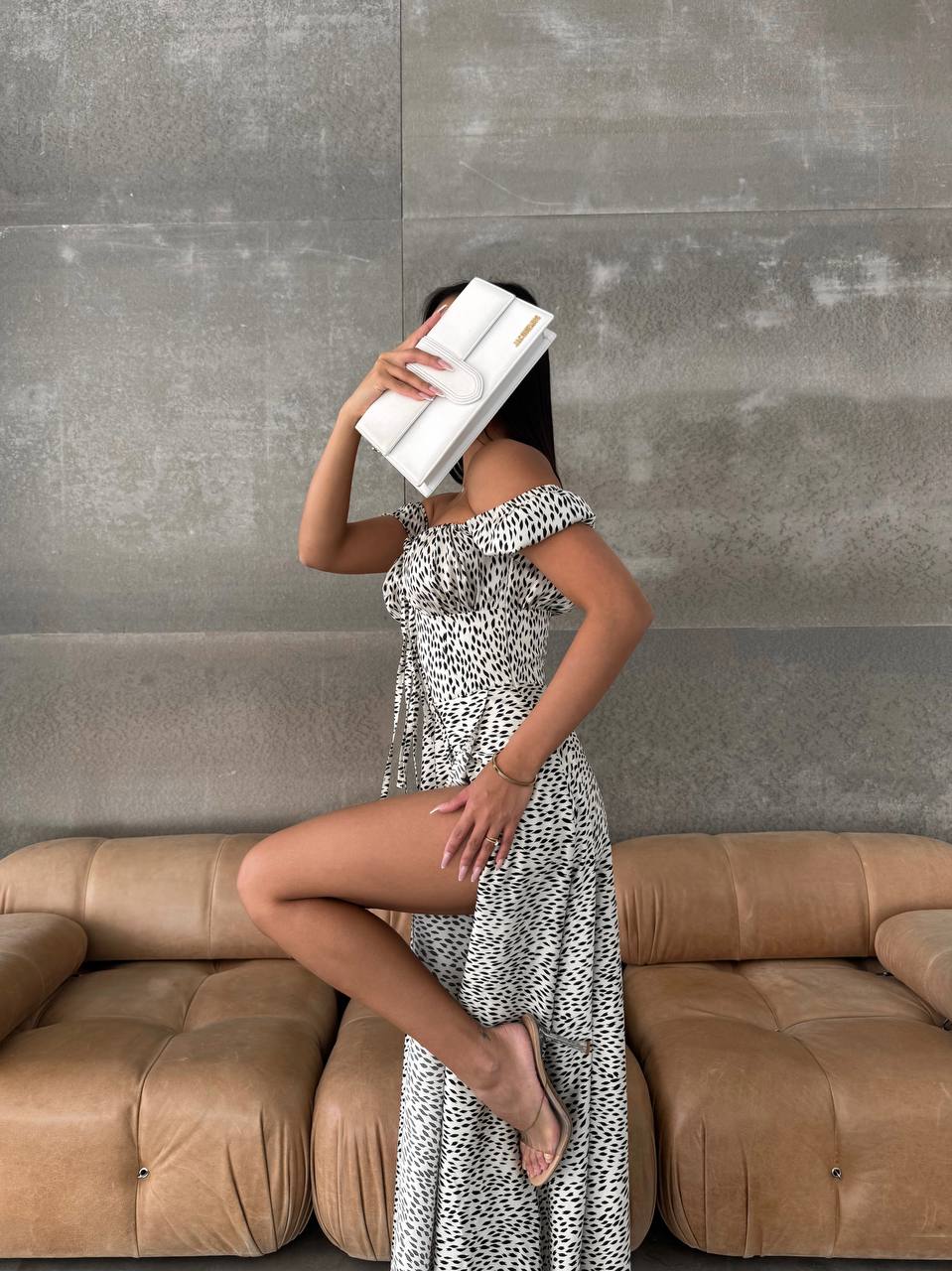 Woman in a patterned dress standing in front of a brown sofa with a gray wall background