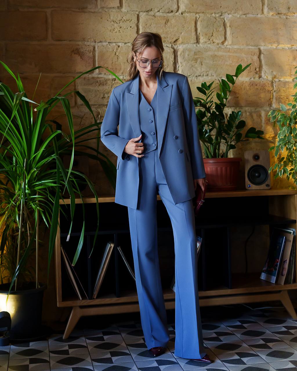 Woman in a blue suit standing in a room with plants and a wooden shelf.