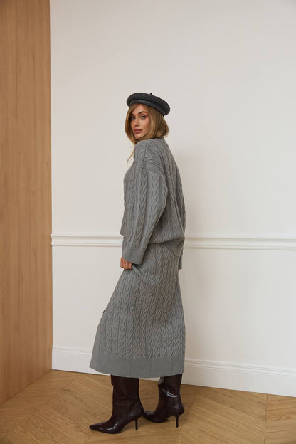 Woman wearing a gray cable knit dress with a matching cardigan and black boots, standing against a white wall.