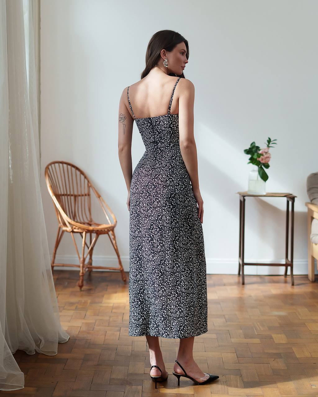 Woman in a patterned dress standing in a room with wooden floor and white walls.