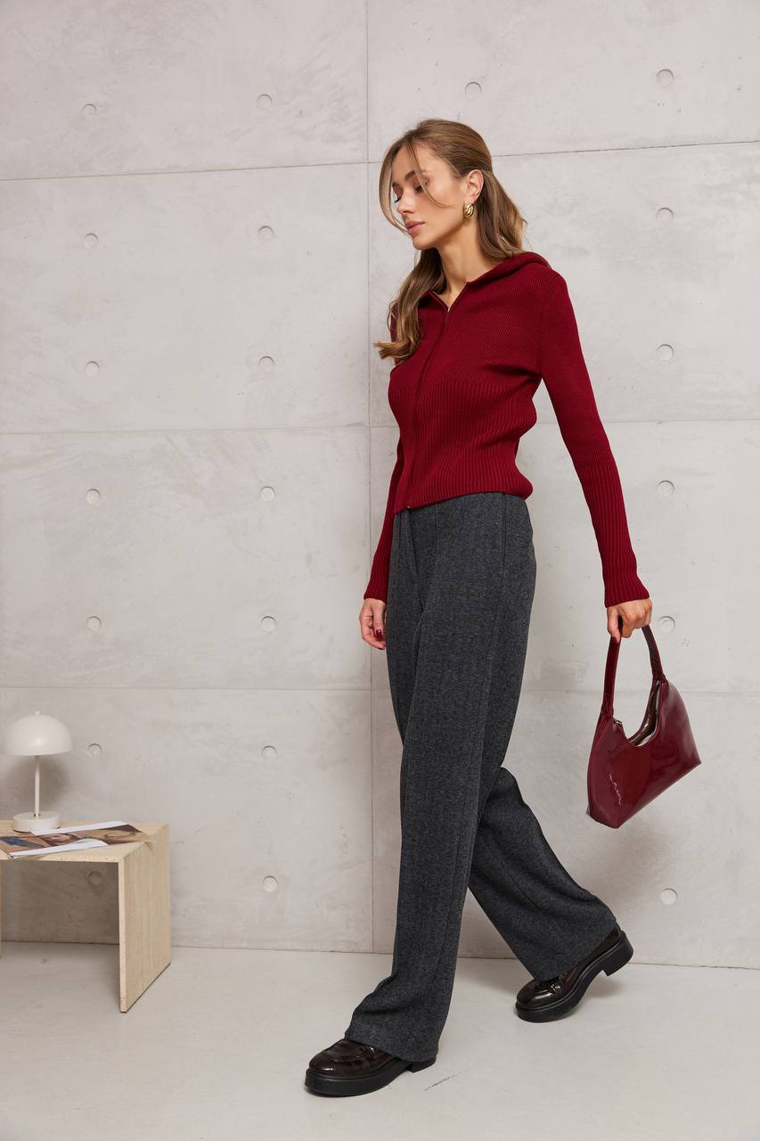 Woman in a red sweater and gray pants holding a red handbag against a light gray wall.