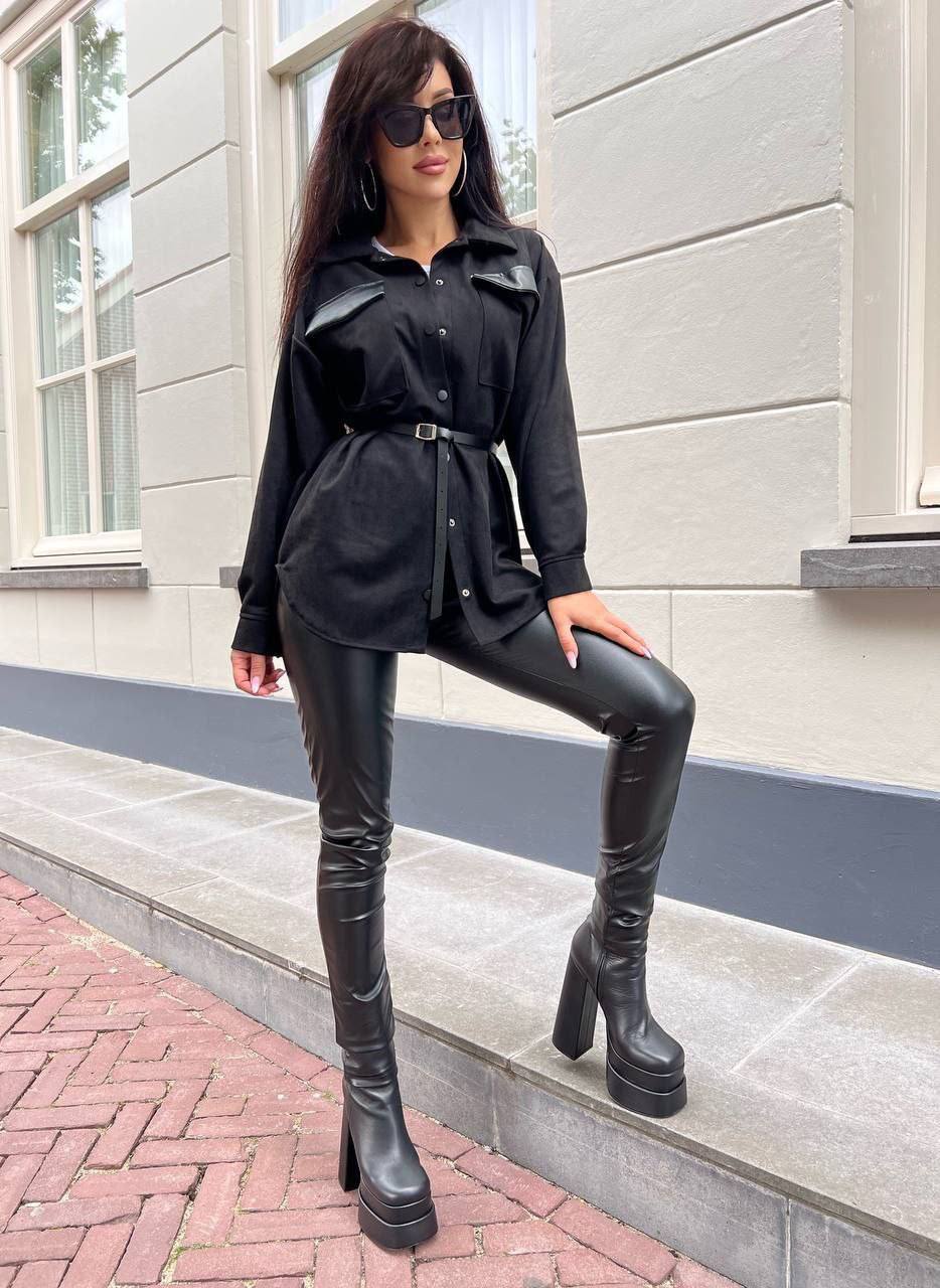 Woman in black outfit with leather pants and boots standing on a sidewalk.