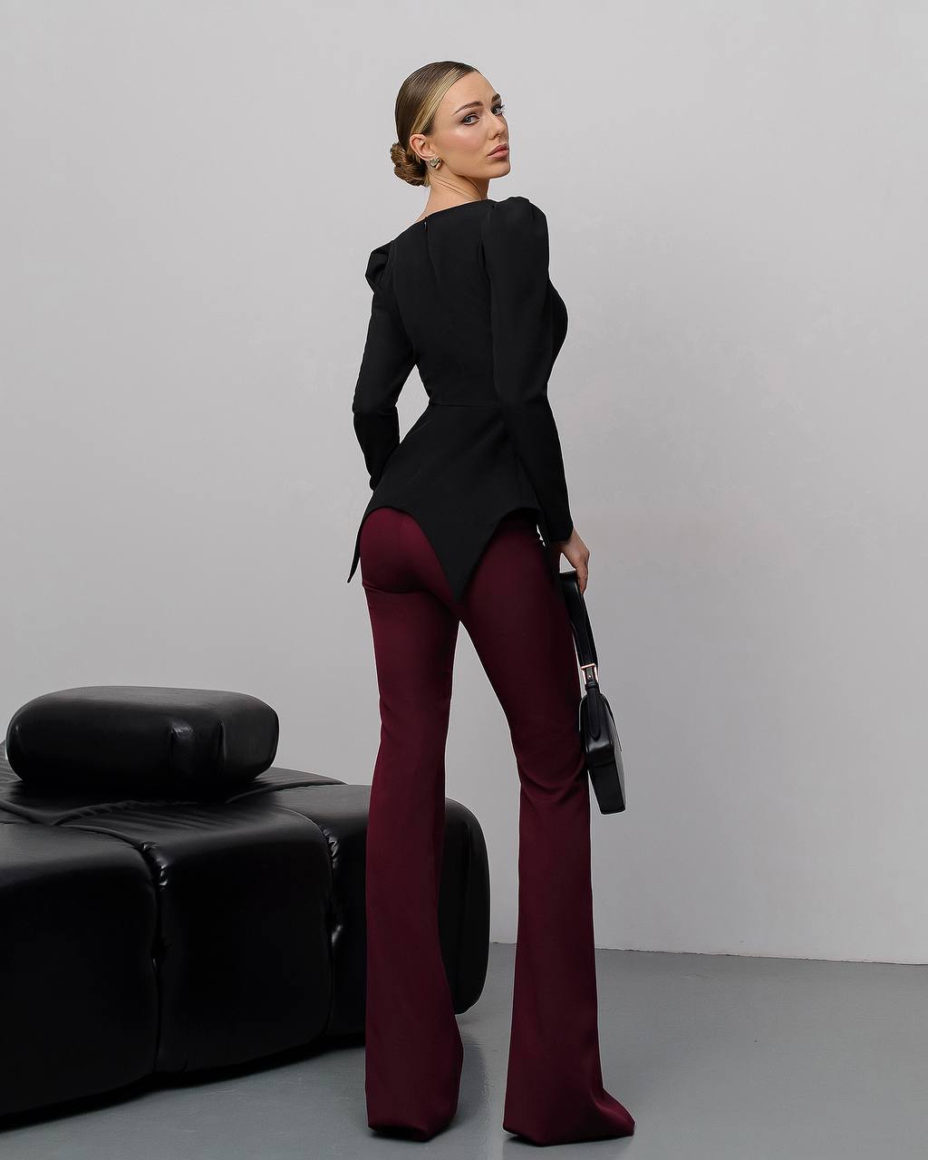 Woman in a black top and burgundy pants standing against a plain background