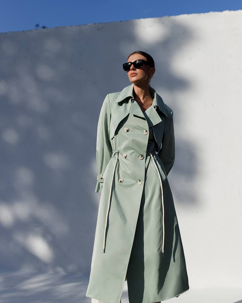 Person wearing a light green trench coat against a white wall with blue sky.