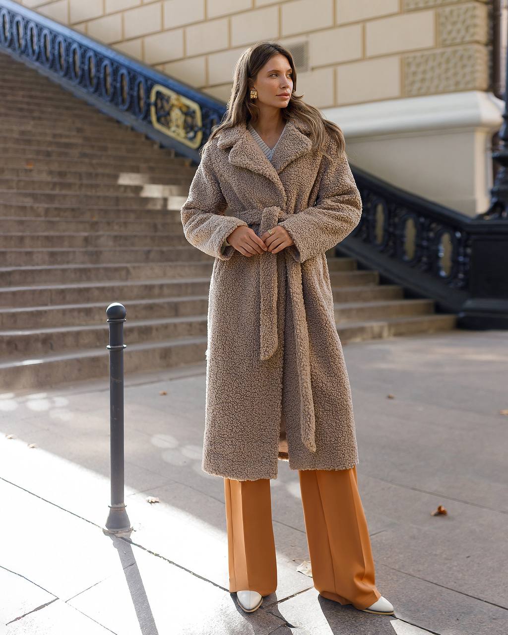 Woman wearing a long beige coat and orange pants standing on a city street.