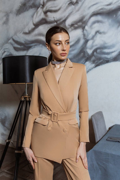 Woman in a beige suit standing in a room with a lamp and decorative wall.