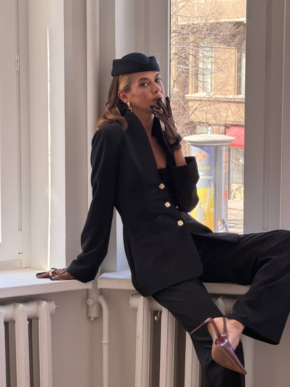 Woman in a black suit sitting by a window with a cityscape view.