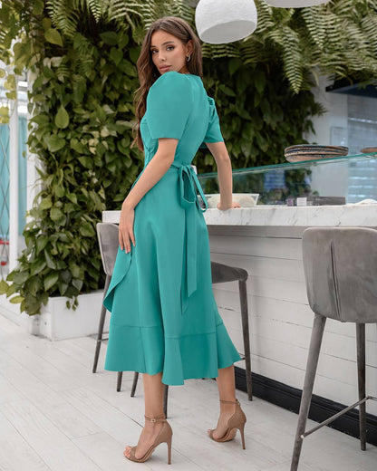 Woman in a teal dress standing in an outdoor setting with greenery and a table.
