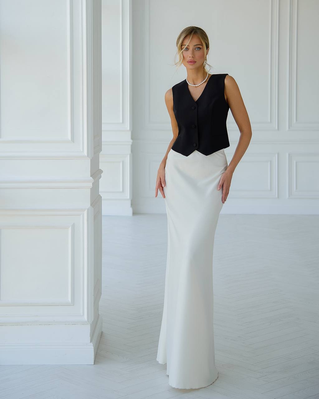 Woman wearing a black sleeveless top and white pants in a white room.
