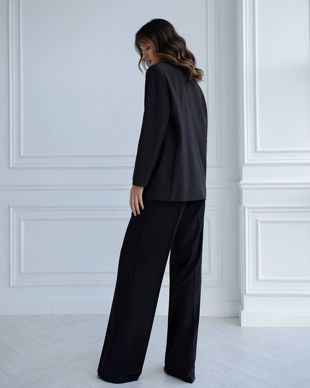 Woman wearing a black suit standing against a white paneled wall.