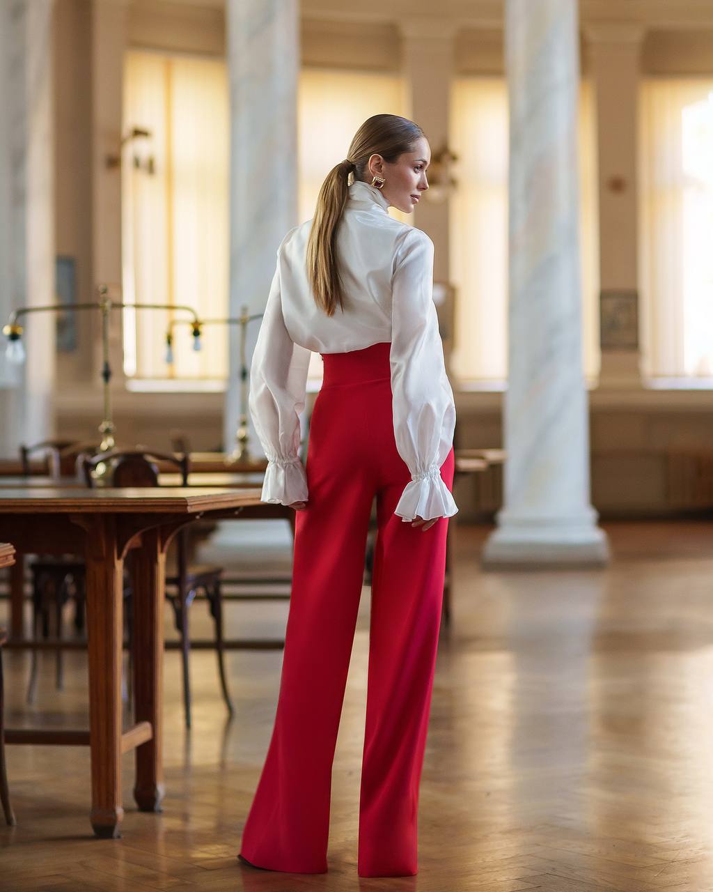 Woman in a white blouse and red pants standing in a large room with columns and wooden tables.