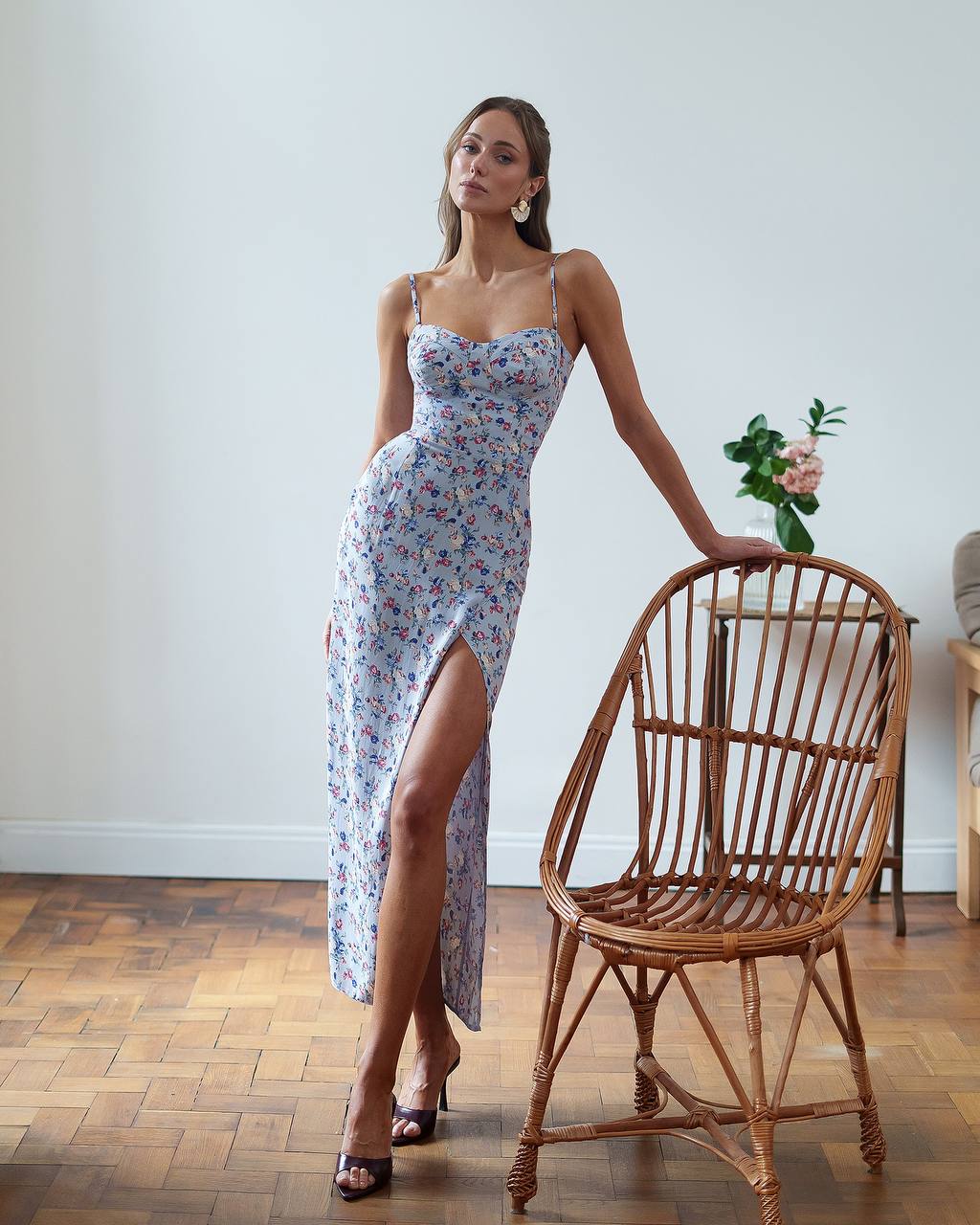 Woman in a floral dress standing next to a wicker chair indoors.