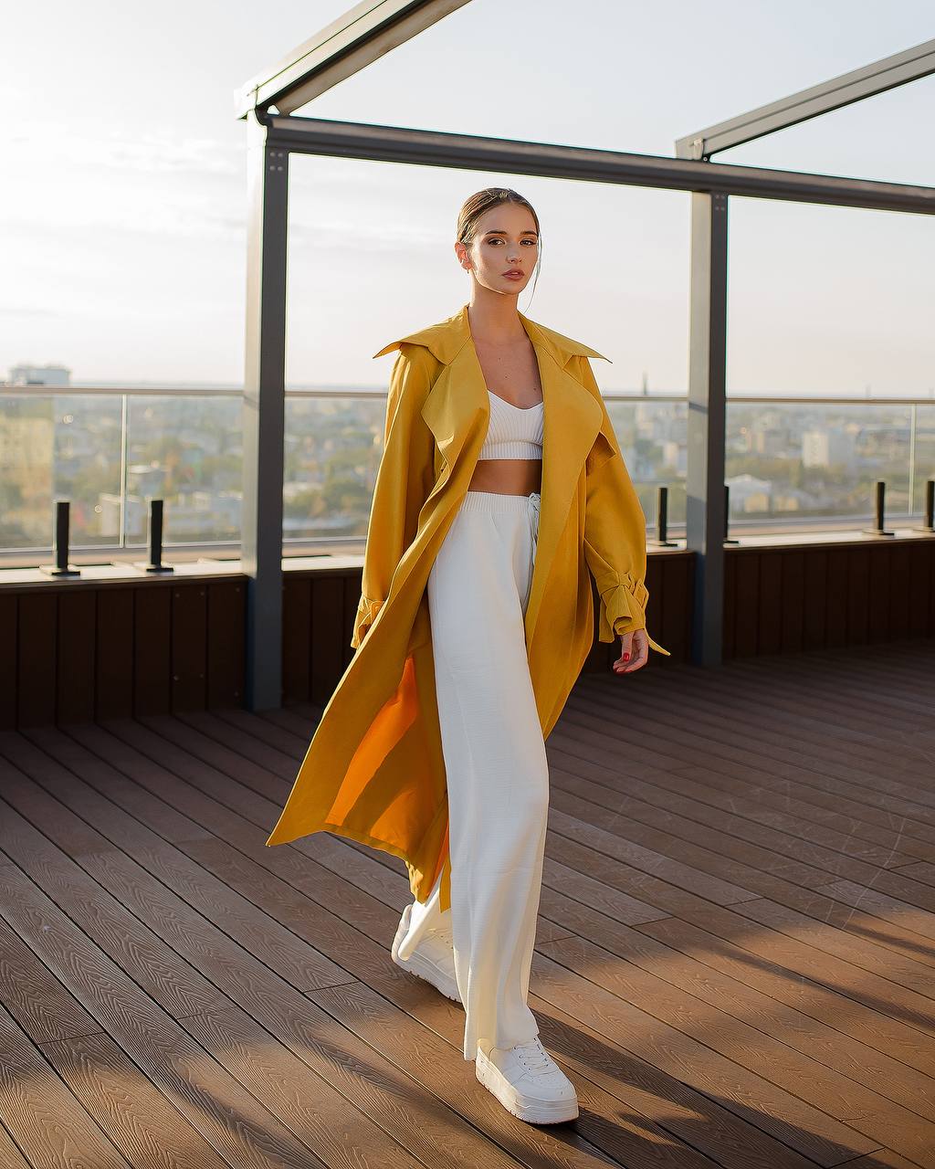 Woman in a yellow trench coat and white outfit on a rooftop with a cityscape view.