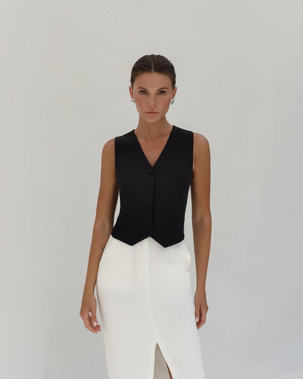 Woman wearing a black sleeveless top and white pants against a plain background