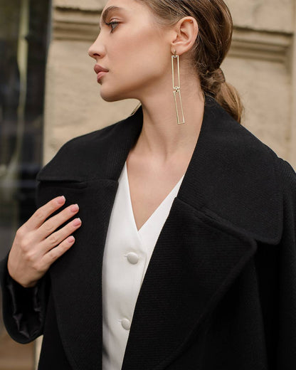 Woman wearing a black coat and white blouse with gold earrings, standing against a blurred background.
