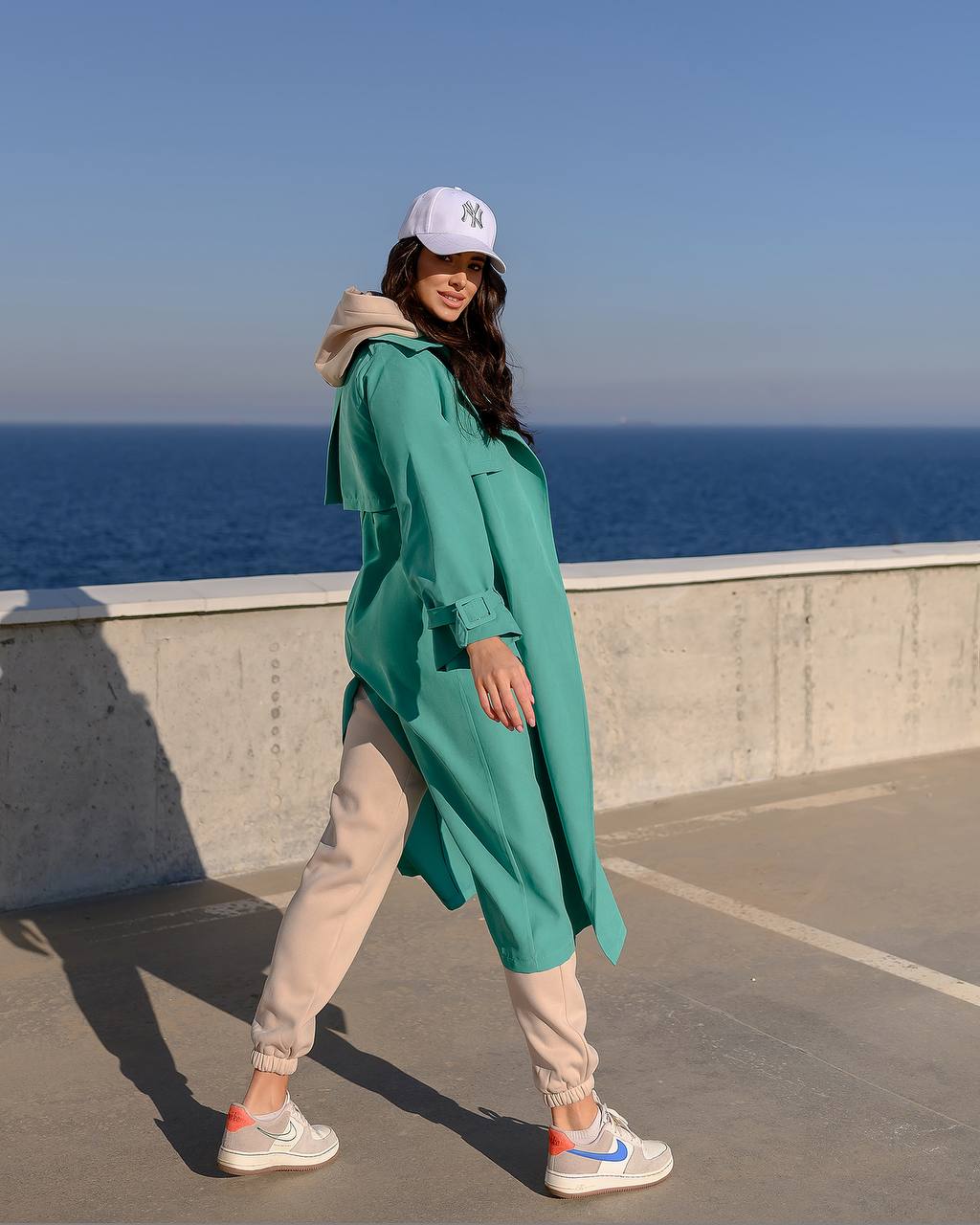 Woman in teal coat and beige pants standing on a rooftop with ocean view