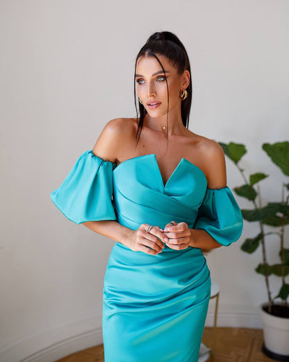 Woman wearing a turquoise off-shoulder dress indoors with a plant in the background