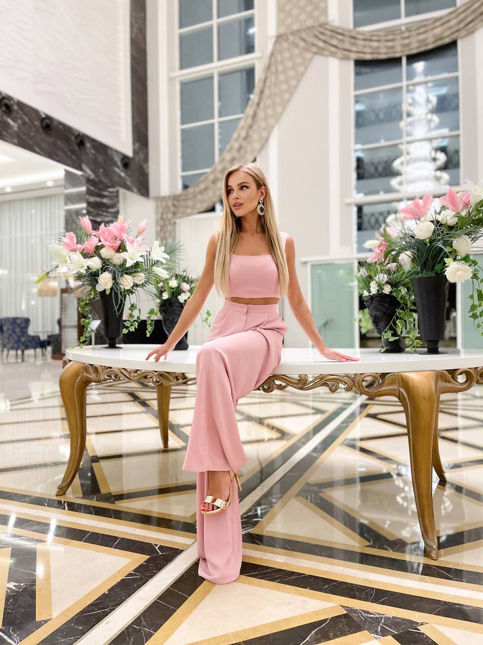Woman in a pink outfit sitting on a marble bench in a luxurious interior setting with flowers and decor.