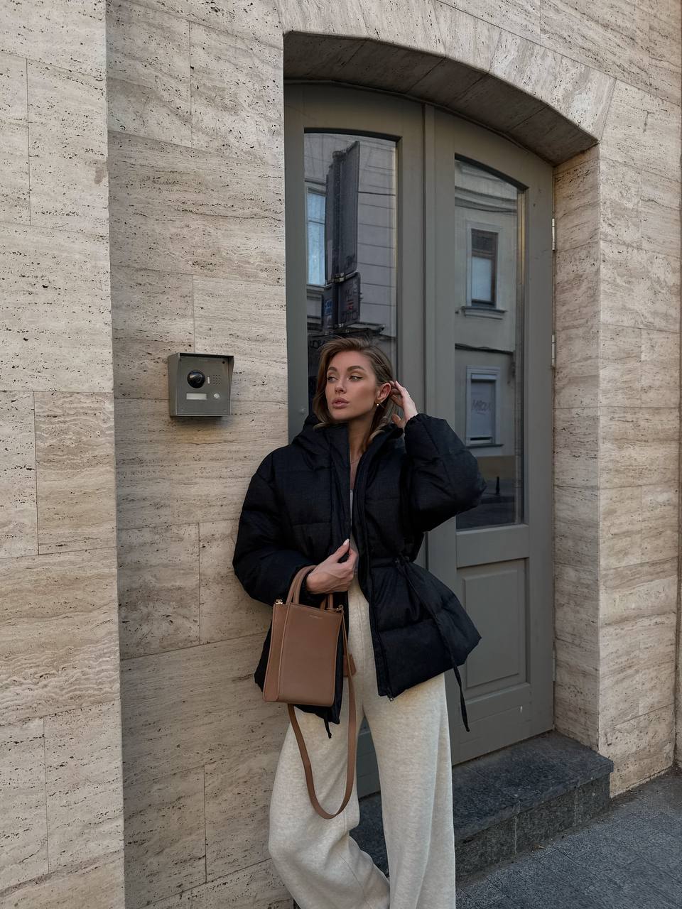 Woman standing in front of a building entrance wearing a black coat and white pants.