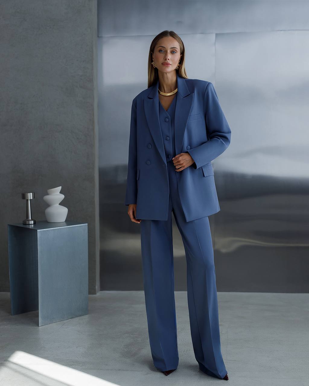 Woman in a blue suit standing in a minimalistic room with gray walls and a small table.